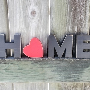 K&ouml;nnte beinhalten: Schwarze Holzbuchstaben, die "HOME" buchstabieren, mit einem roten Herzen zwischen dem "H" und dem "O".
