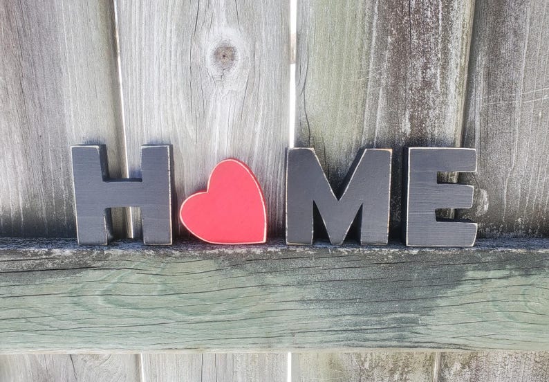 K&ouml;nnte beinhalten: Schwarze Holzbuchstaben, die "HOME" buchstabieren, mit einem roten Herzen zwischen dem "H" und dem "O".