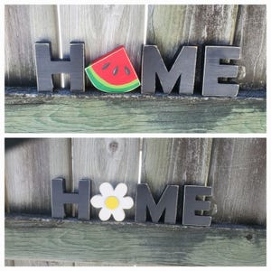 K&ouml;nnte beinhalten: Schwarze Holzbuchstaben, die "HOME" buchstabieren, wobei ein Wassermelonenst&uuml;ck und eine G&auml;nsebl&uuml;mchenbl&uuml;te die "O"s ersetzen.