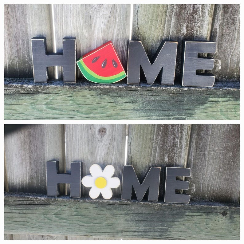 K&ouml;nnte beinhalten: Schwarze Holzbuchstaben, die "HOME" buchstabieren, wobei ein Wassermelonenst&uuml;ck und eine G&auml;nsebl&uuml;mchenbl&uuml;te die "O"s ersetzen.