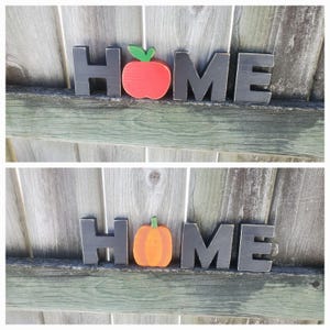 K&ouml;nnte beinhalten: Schwarze Holzbuchstaben, die "HOME" buchstabieren, mit einem roten Apfel und einem orangefarbenen K&uuml;rbis in der Mitte.