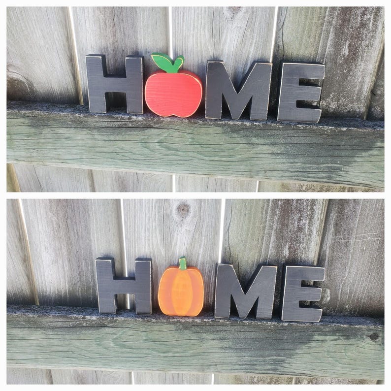 K&ouml;nnte beinhalten: Schwarze Holzbuchstaben, die "HOME" buchstabieren, mit einem roten Apfel und einem orangefarbenen K&uuml;rbis in der Mitte.