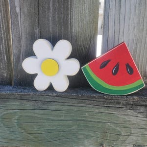K&ouml;nnte beinhalten: Eine wei&szlig;e Margerite mit gelbem Zentrum und ein rotes Wassermelonenst&uuml;ck mit schwarzen Kernen. Beide sind auf Holz gemalt und an einem Holzzaun befestigt.