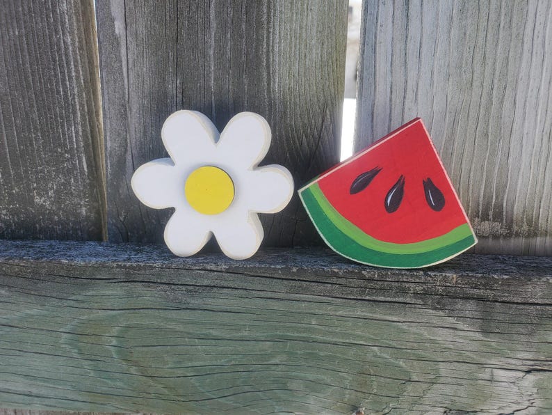 K&ouml;nnte beinhalten: Eine wei&szlig;e Margerite mit gelbem Zentrum und ein rotes Wassermelonenst&uuml;ck mit schwarzen Kernen. Beide sind auf Holz gemalt und an einem Holzzaun befestigt.