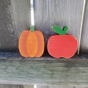 K&ouml;nnte beinhalten: Zwei bemalte Holz-Herbstdekorationen, ein roter Apfel und ein orangefarbener K&uuml;rbis, auf einem Holzgrund.