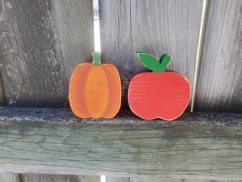 K&ouml;nnte beinhalten: Zwei bemalte Holz-Herbstdekorationen, ein roter Apfel und ein orangefarbener K&uuml;rbis, auf einem Holzgrund.