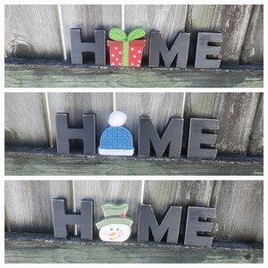 K&ouml;nnte beinhalten: Schwarze Holzbuchstaben, die "HOME" buchstabieren, mit einer anderen weihnachtlichen Dekoration auf jedem Buchstaben. Der erste Buchstabe hat eine rote und gr&uuml;ne Geschenkbox, der zweite einen blauen Strickhut und der dritte einen gr&uuml;nen Schneemann.