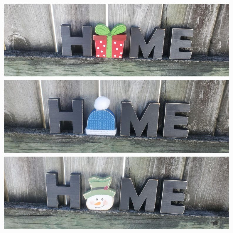 K&ouml;nnte beinhalten: Schwarze Holzbuchstaben, die "HOME" buchstabieren, mit einer anderen weihnachtlichen Dekoration auf jedem Buchstaben. Der erste Buchstabe hat eine rote und gr&uuml;ne Geschenkbox, der zweite einen blauen Strickhut und der dritte einen gr&uuml;nen Schneemann.