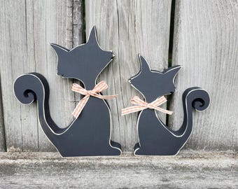 May include: Two black wooden cat silhouettes with pink and white gingham bows. The larger cat is on the left and the smaller cat is on the right. The cats are facing each other.