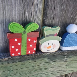 K&ouml;nnte beinhalten: Drei h&ouml;lzerne Weihnachtsornamente: eine rote und wei&szlig;e Geschenkbox mit wei&szlig;en Punkten und einer gr&uuml;nen Schleife, ein Schneemann mit gr&uuml;nem Hut und einer Karottennase und eine blaue Strickm&uuml;tze mit einem wei&szlig;en Bommel.