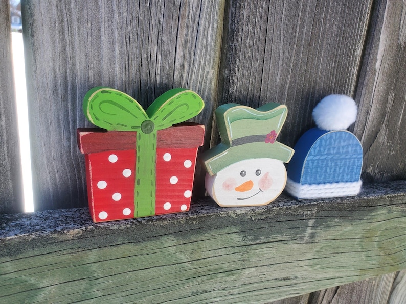 K&ouml;nnte beinhalten: Drei h&ouml;lzerne Weihnachtsornamente: eine rote und wei&szlig;e Geschenkbox mit wei&szlig;en Punkten und einer gr&uuml;nen Schleife, ein Schneemann mit gr&uuml;nem Hut und einer Karottennase und eine blaue Strickm&uuml;tze mit einem wei&szlig;en Bommel.