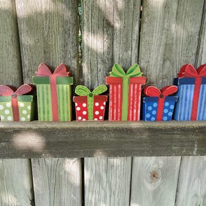 May include: A collection of six wooden gift boxes with colorful designs. The boxes feature red, green, and blue hues, with polka dots, stripes, and bows. They are arranged on a wooden surface against a weathered wooden fence.