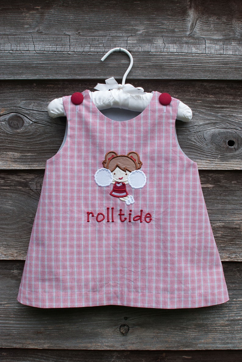 May include: A sleeveless red and white plaid dress with a cheerleader applique and the embroidered text "rolltide". The dress has red buttons on the shoulders and is hanging on a white hanger.