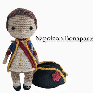 K&ouml;nnte beinhalten: Geh&auml;kelte Amigurumi-Puppe von Napoleon Bonaparte, die eine wei&szlig;e, rote und blaue Uniform mit einem schwarzen geh&auml;kelten Hut tr&auml;gt. Die Puppe steht auf einem wei&szlig;en Hintergrund.
