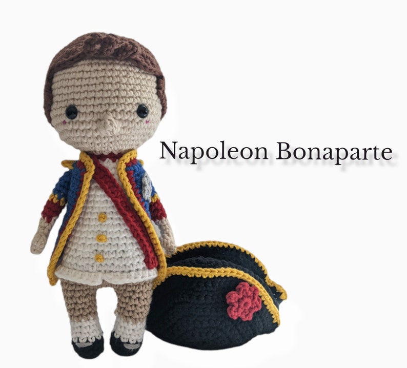 K&ouml;nnte beinhalten: Geh&auml;kelte Amigurumi-Puppe von Napoleon Bonaparte, die eine wei&szlig;e, rote und blaue Uniform mit einem schwarzen geh&auml;kelten Hut tr&auml;gt. Die Puppe steht auf einem wei&szlig;en Hintergrund.