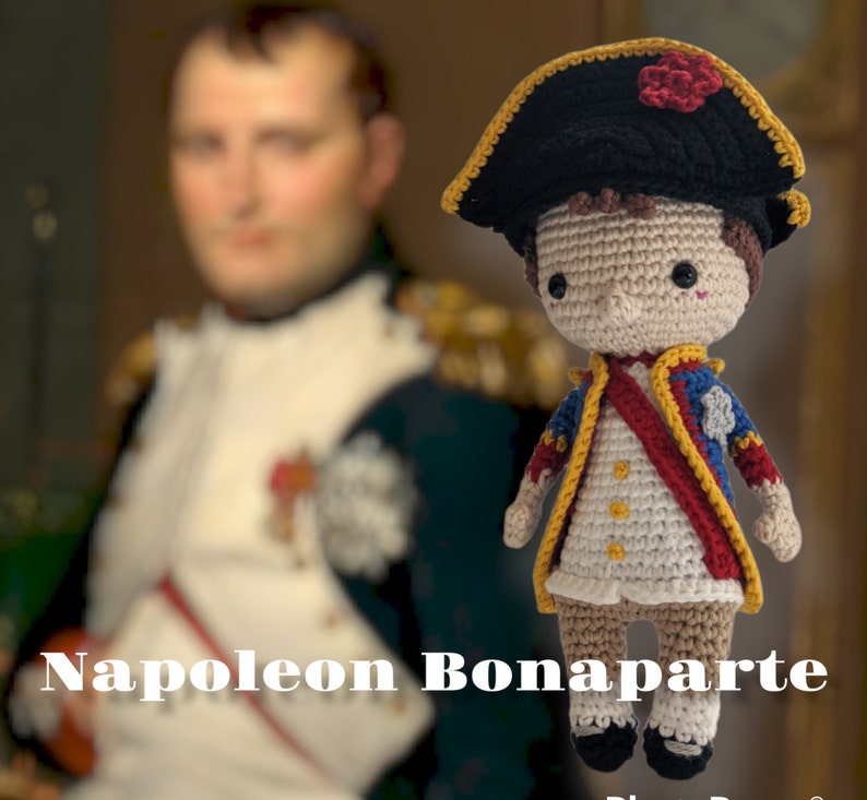 K&ouml;nnte beinhalten: Eine geh&auml;kelte Puppe von Napoleon Bonaparte, die eine blaue und rote Uniform mit goldenen Verzierungen und einen schwarzen Hut mit einer roten Blume tr&auml;gt. Die Puppe steht auf einer wei&szlig;en Oberfl&auml;che mit dem Text "Napoleon Bonaparte" in schwarzen Buchstaben.