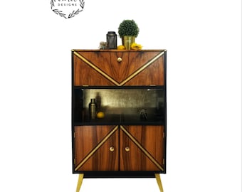 May include: A mid-century modern bar cabinet with gold accents and black and wood veneer doors. The cabinet has a glass shelf and gold legs.