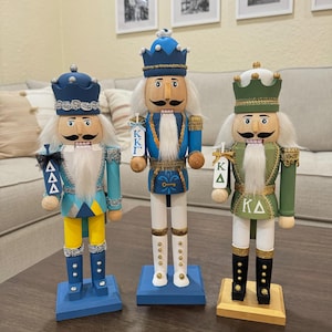 May include: Three wooden nutcracker figurines, each with a different color scheme and a different Greek letter fraternity symbol. The nutcracker on the left is blue and yellow with the letters "ΔΔΔ". The nutcracker in the middle is blue and white with the letters "ΚΚΓ". The nutcracker on the right is green and white with the letters "ΚΔ".