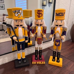 Puede incluir: Tres cascanueces de madera con sombreros y uniformes amarillos. Uno sostiene un palo de hockey, otro tiene un tambor y el tercero tiene baquetas. Cada cascanueces tiene un logotipo diferente en su sombrero y su base.