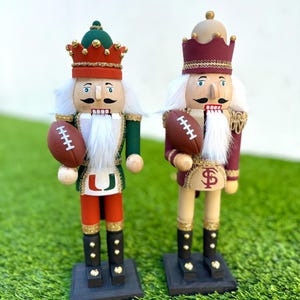 May include: Two wooden nutcrackers, each holding a football. One is dressed in green, orange, and red, the other in maroon and gold. Both have white beards, crowns, and black boots, standing on a green surface.