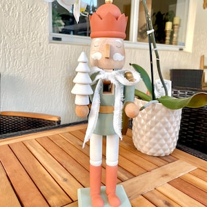 Puede incluir: Una figura de cascanueces de madera con un atuendo verde y blanco, un sombrero rojo y una barba blanca. El cascanueces sostiene un pequeño árbol de Navidad blanco.