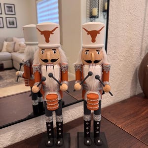 Puede incluir: Dos cascanueces de madera con forma de soldados de juguete. Llevan sombreros blancos con un logotipo de cuerno largo naranja quemado, chaquetas naranjas y botas negras. Cada uno sostiene baquetas y tiene un pequeño tambor naranja.