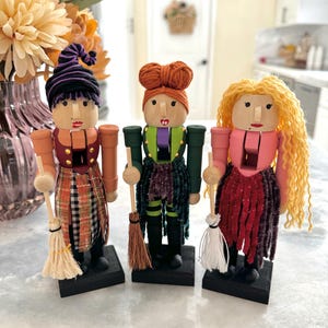 Op de afbeelding: Drie houten notenkraker-figuurtjes verkleed als heksen. Elke heks heeft een andere gekleurde jurk en haar. De heks links heeft een paarse hoed en een geruite rok. De heks in het midden heeft een groene jurk en een groen-zwarte gestreepte rok. De heks rechts heeft een roze jurk en een rode rok met glitter. Elke heks houdt een bezem.