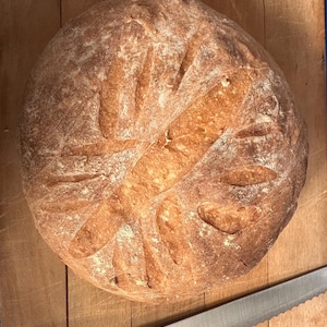 Puede incluir: Un pan redondo recién horneado, con una corteza dorada y cortes decorativos. El pan está sobre una tabla de cortar de madera, con un cuchillo de sierra cerca. Aspecto rústico y casero.