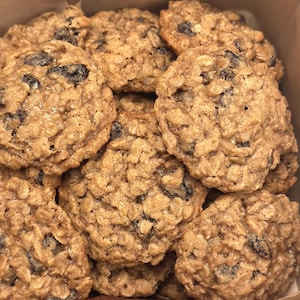 Puede incluir: Primer plano de una caja llena de galletas de avena y pasas. Las galletas son de color marrón dorado, con avena visible y pasas oscuras. Las galletas son redondas y tienen una superficie ligeramente texturizada.