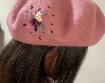 Op de afbeelding: Een roze wollen baret met een geborduurd bloemenmotief. De bloemen zijn geborduurd in tinten paars, wit en groen.