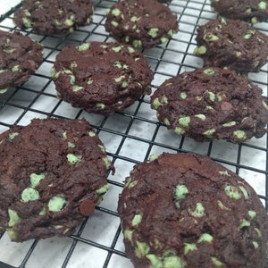 May include: A close-up of a cooling rack with freshly baked chocolate chip cookies with green mint chips. The cookies are dark brown and have a slightly crispy texture.