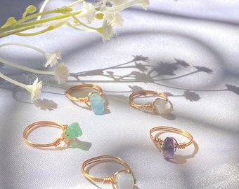 May include: Five gold rings with different coloured gemstones. The rings are on a white fabric background. The gemstones are aquamarine, green, white, amethyst, and white.