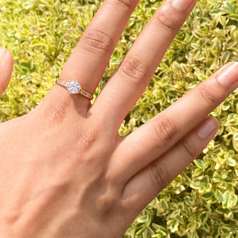 May include: A gold ring with a large, clear, round gemstone surrounded by smaller gemstones. The ring is on a finger.