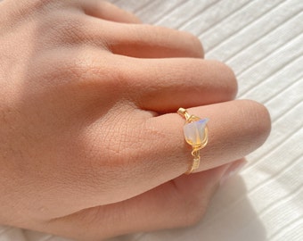May include: A gold-coloured wire-wrapped ring with a small, iridescent, rectangular gemstone. The ring is on a finger, highlighting the delicate craftsmanship and the stone's subtle colour variations.