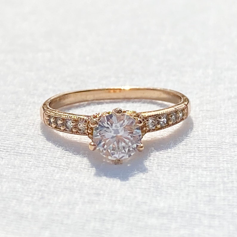 May include: A rose gold engagement ring with a single round-cut diamond centre stone and a band of smaller diamonds.