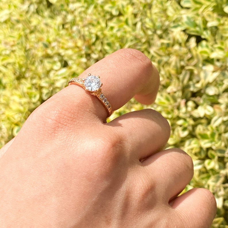 May include: A gold ring with a large, clear, round gemstone surrounded by smaller gemstones. The ring is on a finger.
