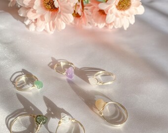 May include: Five gold rings with different coloured stones. The stones are green, purple, white, orange, and clear. The rings are on a white surface.