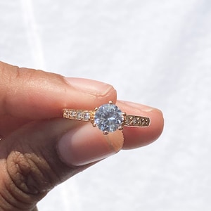 May include: A gold ring featuring a large, clear, round gemstone surrounded by smaller, clear gemstones. The ring is set against a plain white background.