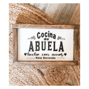 May include: A wooden sign with the text "Cocina de Abuela - hecho con amor - Nietos Bienvenidos" in black lettering on a white background. The sign is framed in a rustic brown wood frame.