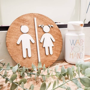 May include: A round wooden sign with a white silhouette of a boy and girl on either side of a dividing line. The sign reads "WASH YOUR HANDS" in colorful letters.