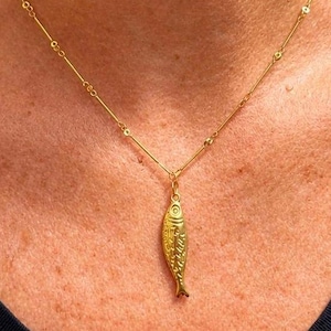 May include: A gold-toned necklace featuring a delicate chain with small, evenly spaced metal beads and a central pendant shaped like a stylised fish with detailed scale engravings. The necklace rests against a plain dark-coloured fabric neckline.