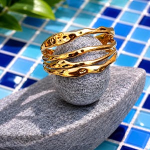 May include: A set of three gold-toned bangles featuring a wavy, irregular, and hammered texture. The bracelets have a high-shine, reflective finish and are stacked together, creating a layered appearance with organic, fluid curves along the edges.
