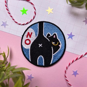 Könnte beinhalten: Ein runder, blauer und weißer gestickter Aufnäher mit einer schwarzen Katze mit weißem Schwanz und dem Wort "NO" in roten und weißen Buchstaben.