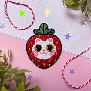 Könnte beinhalten: Ein gestickter Patch in Form einer roten Erdbeere mit einem weißen Katzenkopf darin. Die Katze hat schwarze Augen und eine rosa Nase.