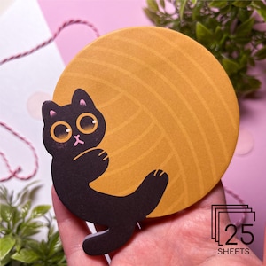 Könnte beinhalten: Eine schwarze Katze mit gelben Augen liegt auf einem großen, gelben Wollknäuel. Das Wollknäuel hat eine strukturierte Oberfläche und ist ein beliebtes Bastelmaterial. Das Bild enthält den Text "25 Blätter".