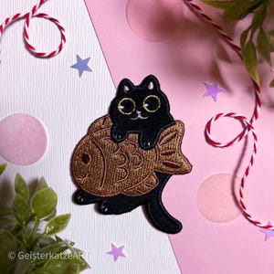 Könnte beinhalten: Ein schwarzer Katzenaufnäher mit goldumrandeten Augen sitzt auf einem braunen, fischförmigen Gebäck. Der Aufnäher befindet sich auf einem rosa-weißen Hintergrund mit dekorativen Sternen und Schnur. Die Katze hat ein niedliches, cartoonhaftes Design.