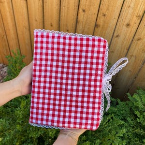 May include: A red and white gingham pattern journal with white lace trim and a matching ribbon tie. The journal is held open, showcasing the quilted fabric design. The cover is a classic, country-style design.