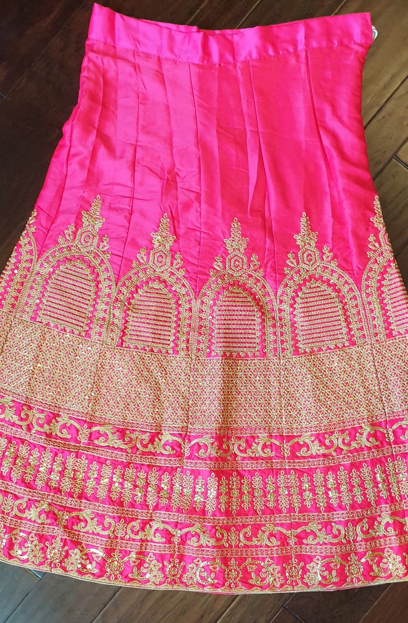 May include: A hot pink skirt with gold embroidery. The skirt has a pleated design and a wide waistband. The embroidery features intricate floral patterns.