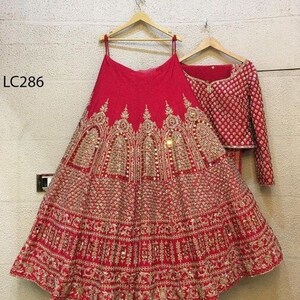 May include: A red bridal lehenga with gold embroidery and sequins. The skirt is layered and has a scalloped hem. The blouse is short-sleeved and has a high neckline. The dupatta is embroidered and has a fringe trim. The outfit is labeled "LC286".