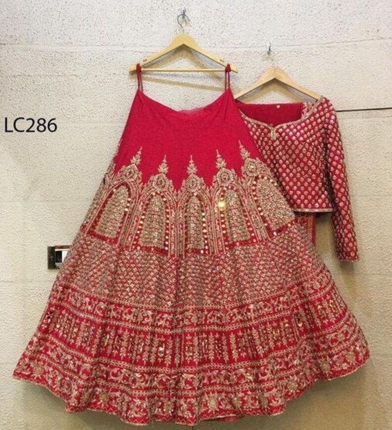 May include: A red bridal lehenga with gold embroidery and sequins. The skirt is layered and has a scalloped hem. The blouse is short-sleeved and has a high neckline. The dupatta is embroidered and has a fringe trim. The outfit is labeled "LC286".